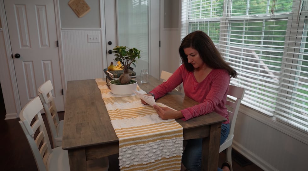 A woman is pictured sitting at a kitchen table, looking down at a piece of paper.